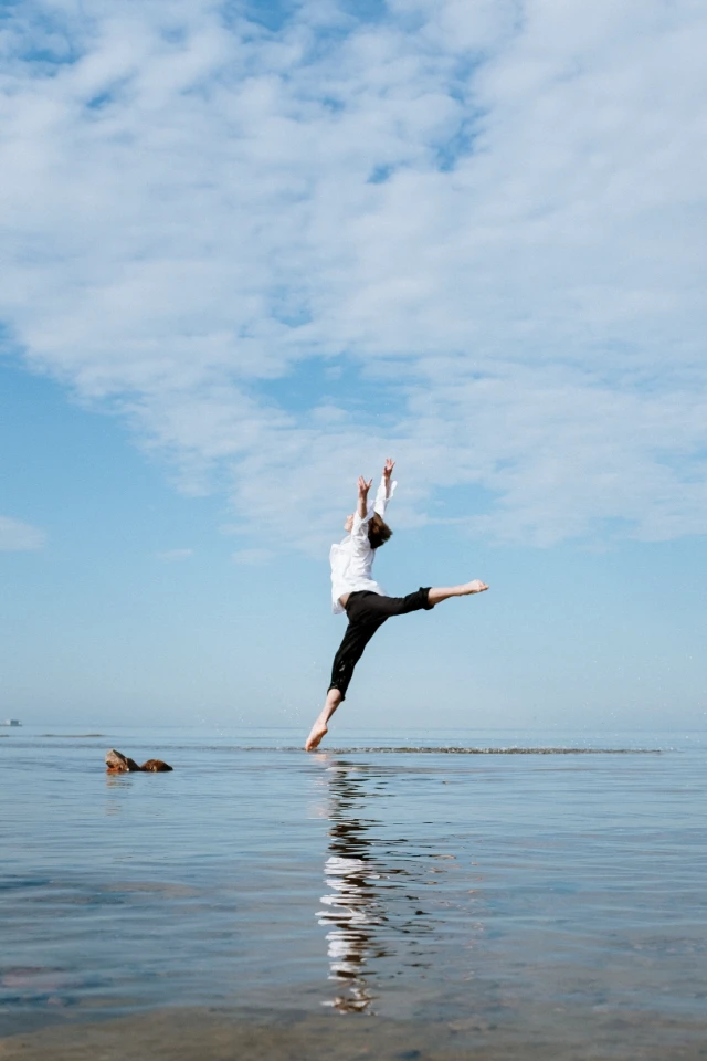 Un danseur en tenue blanche et pantalon noir saute au-dessus de l'eau calme, reflétant sa silhouette sous un ciel bleu et nuageux.