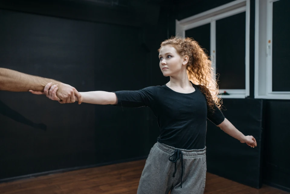 Une jeune femme rousse avec une couette, vêtue d'un t-shirt noir et d'un pantalon de survêtement gris, prend la main d'un partenaire dans une salle de danse.