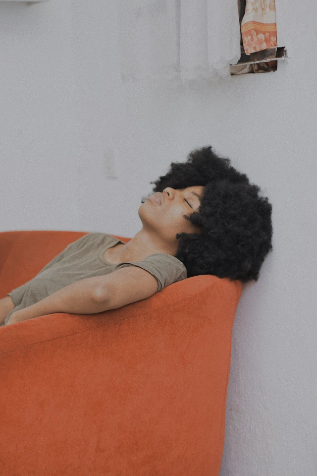 Une femme à la chevelure afro repose le visage incliné en arrière, les yeux fermés, sur un canapé orange vif.
