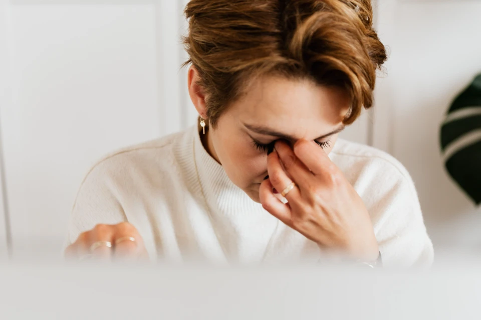 Une femme aux cheveux courts et châtains se frotte les yeux avec les doigts, le visage marqué par la fatigue ou le stress. Elle porte un pull blanc et des bijoux discrets.