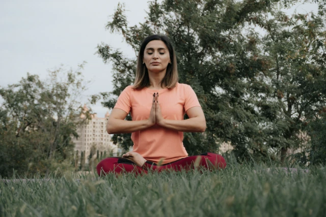 Une femme aux yeux fermés, assise en tailleur sur l'herbe, pratique la méditation avec les mains jointes devant sa poitrine.
