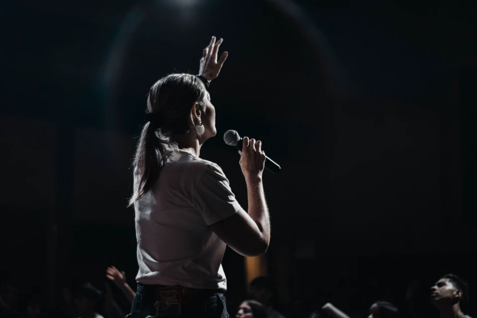 Une femme parle dans un microphone, la main levée, sur une scène éclairée par un projecteur. Le public est flou en arrière-plan.