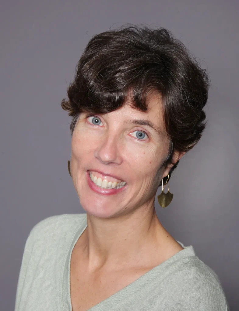Portrait d'une femme aux cheveux bruns courts et aux yeux bleus, souriant avec une chemise vert pâle et des boucles d'oreilles en forme d'éventail.