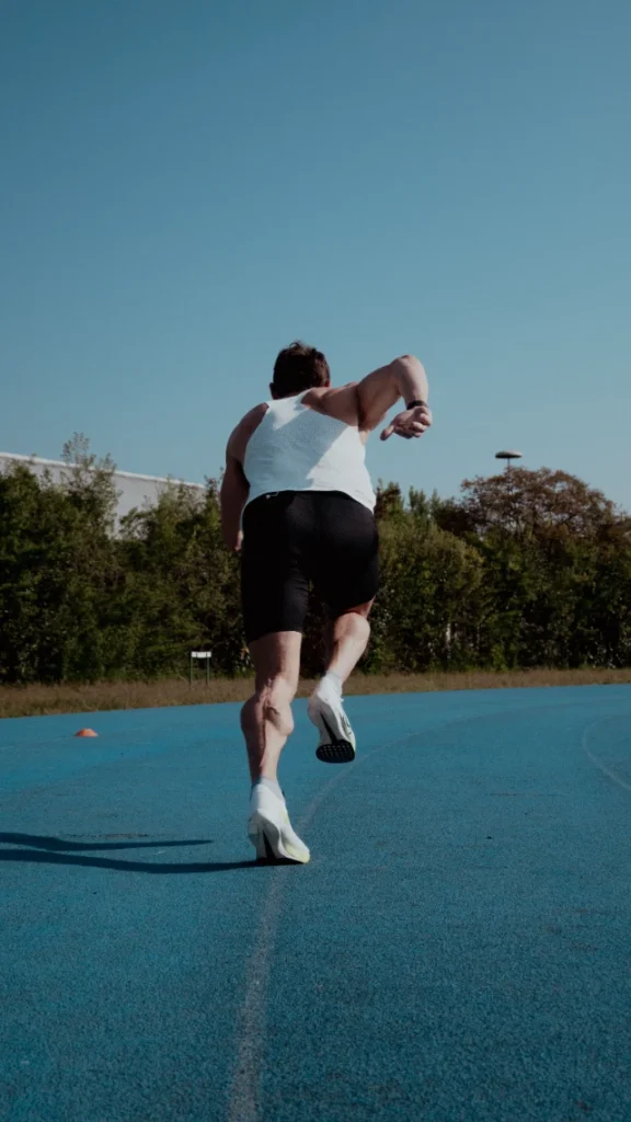 Vue arrière d'un homme musclé courant sur une piste d'athlétisme bleue, portant un débardeur blanc et un short noir.