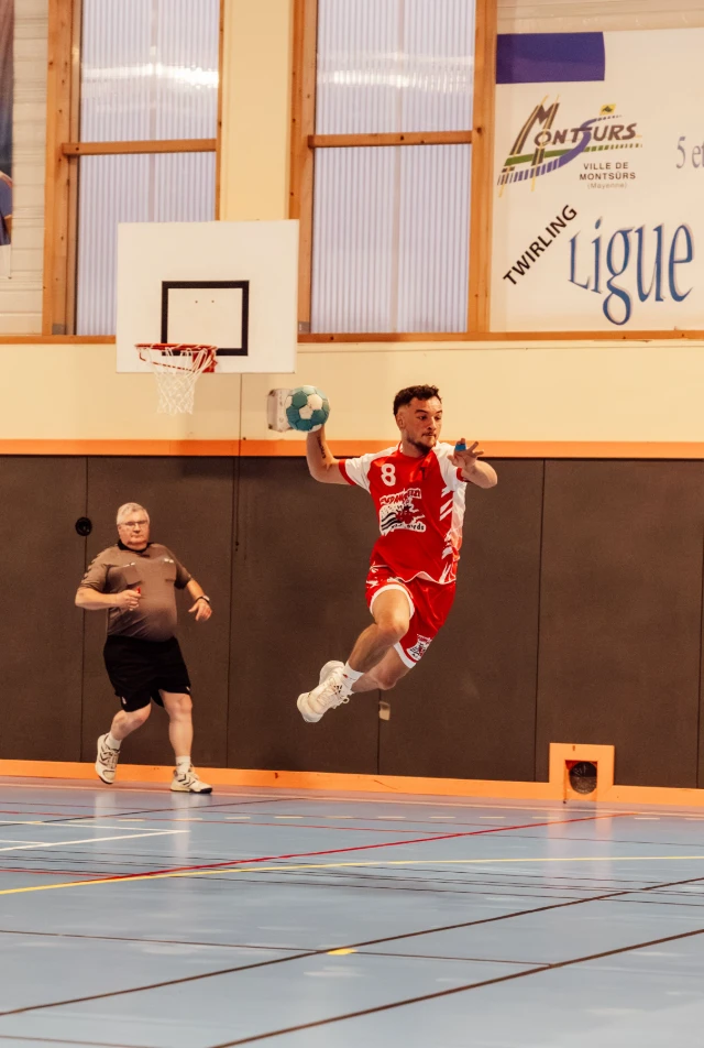 Un joueur de handball en l'air, portant un maillot rouge et blanc, prêt à lancer le ballon. Un autre joueur observe en arrière-plan.