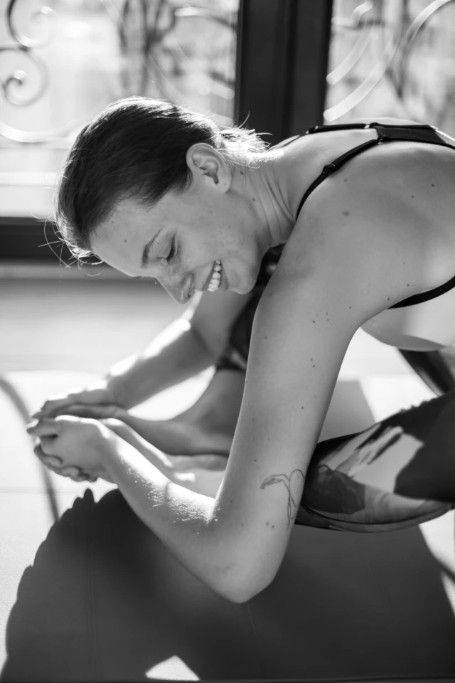 Femme souriante faisant un étirement avant en yoga, se penchant en avant avec les mains jointes sur ses pieds.